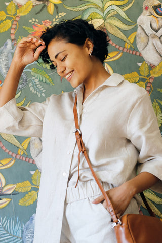 Woman in a light linen top and linen bottoms with a brown bag against a colourful floral wallpaper background.