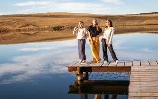 Three women standing on a wooden dock by a lake with a scenic background, all wearing De Tiamo luxury linen tops, pants, jackets and skirts respectively. 