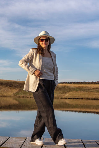 Woman standing on a wooden dock by a body of water, wearing a wide-brimmed hat, sunglasses, black linen pants, a light linen tank top, and a light linen jacket,